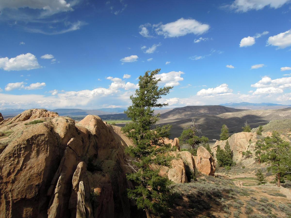 A landscape view featuring rocky formations and trees under a blue sky with scattered clouds. The scene includes rolling hills in the background, creating a sense of depth and natural beauty. Hartman Rocks mountain bike trail.