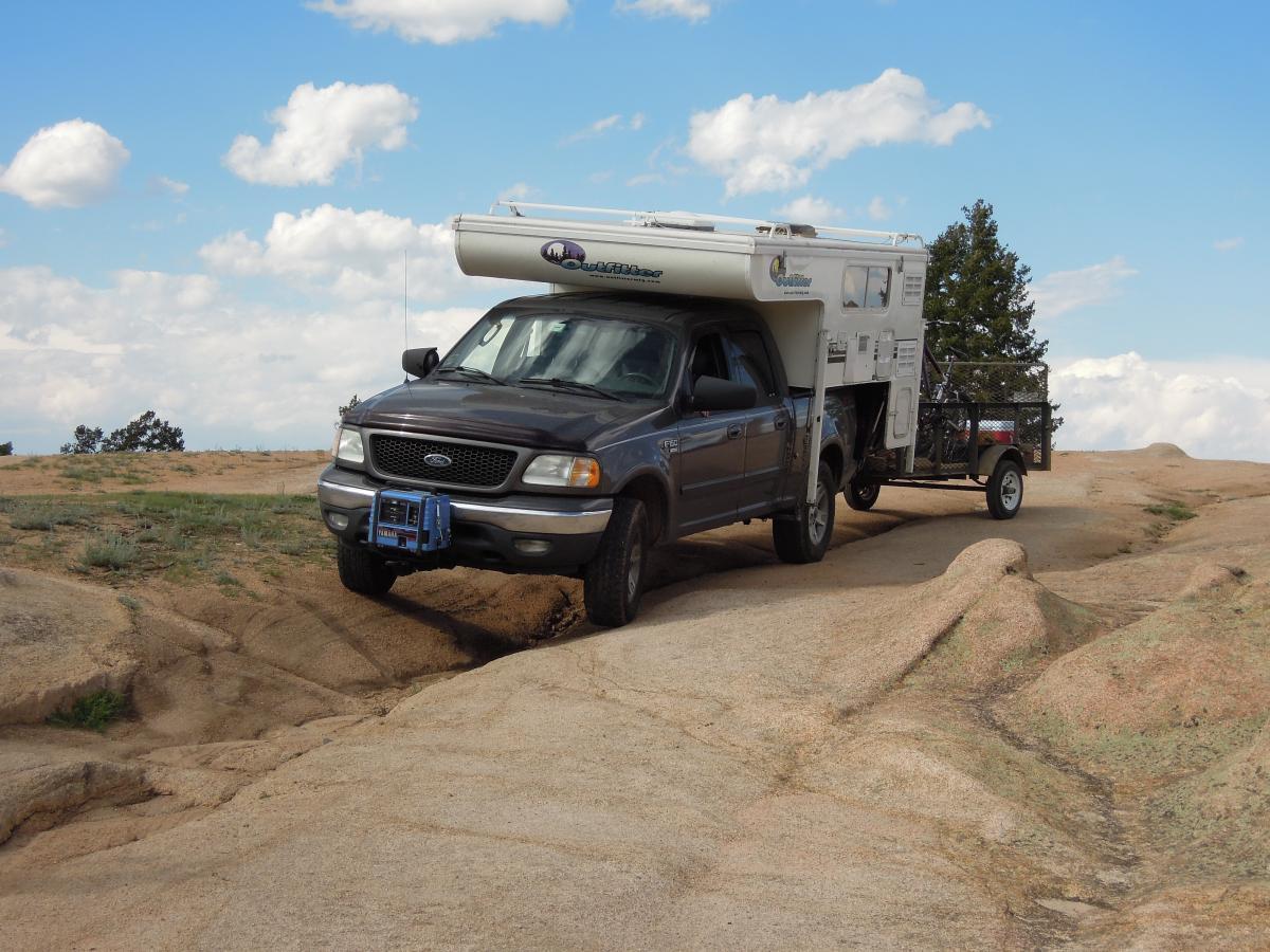 A gray pickup truck with a camper shell is parked on a rocky terrain, with a small trailer attached. The background features a blue sky with fluffy white clouds and sparse trees. The ground is uneven, showcasing the rugged landscape typical of off-road settings. Hartman Rocks mountain bike trail.