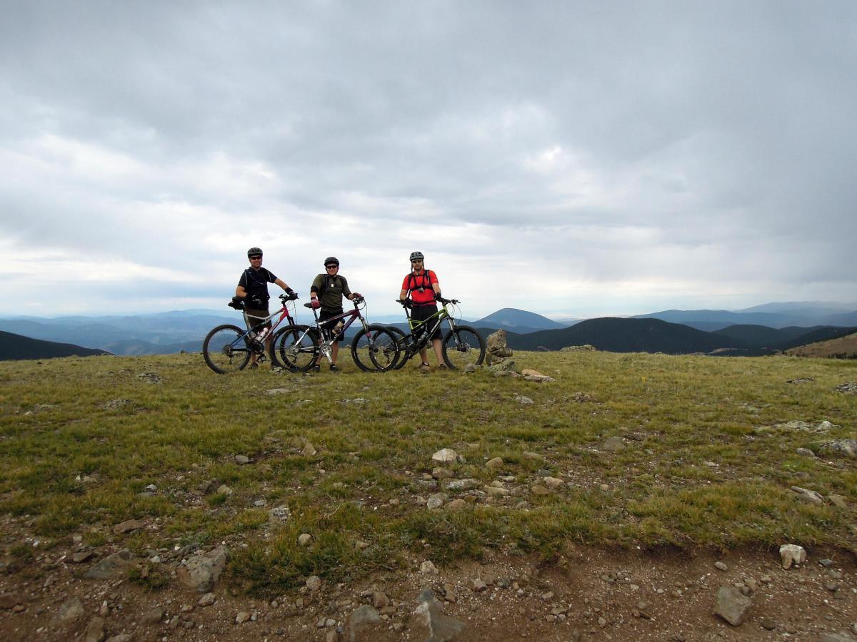 Three mountain bikers pose with their bicycles on a grassy plateau surrounded by mountains under a cloudy sky. The landscape features rolling hills in the background, and the bikers are dressed in cycling gear with helmets. Monarch Crest Trail mountain bike trail.