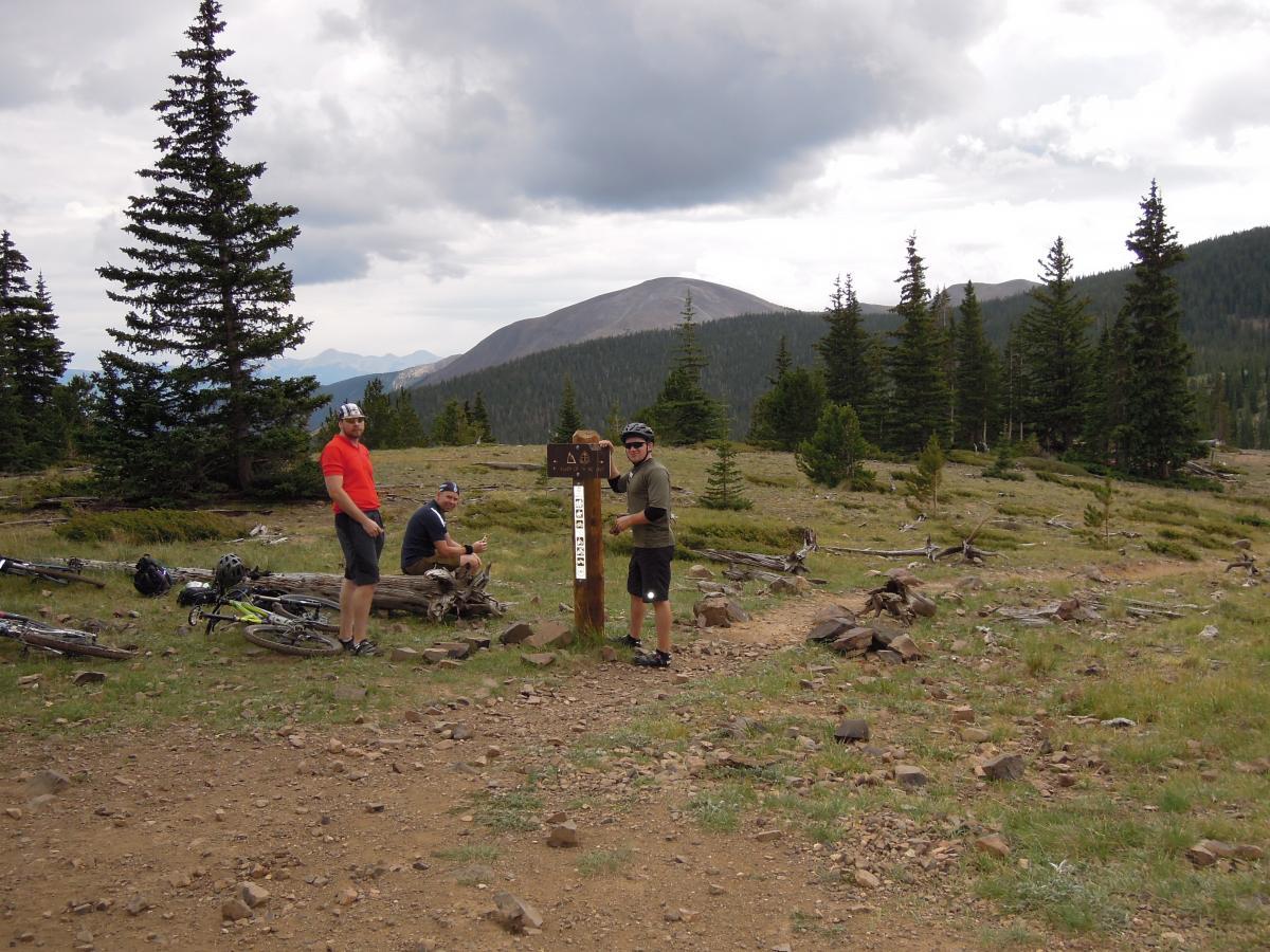 A group of three mountain bikers pauses at a trail sign in a mountainous area, surrounded by pine trees and rocky terrain. Two riders stand beside the sign, one wearing a green shirt and helmet, while the other is in a red shirt. The third biker sits on a log nearby, resting. Bicycles are leaning against the log, with rugged mountains visible in the background under a cloudy sky. Monarch Crest Trail mountain bike trail.