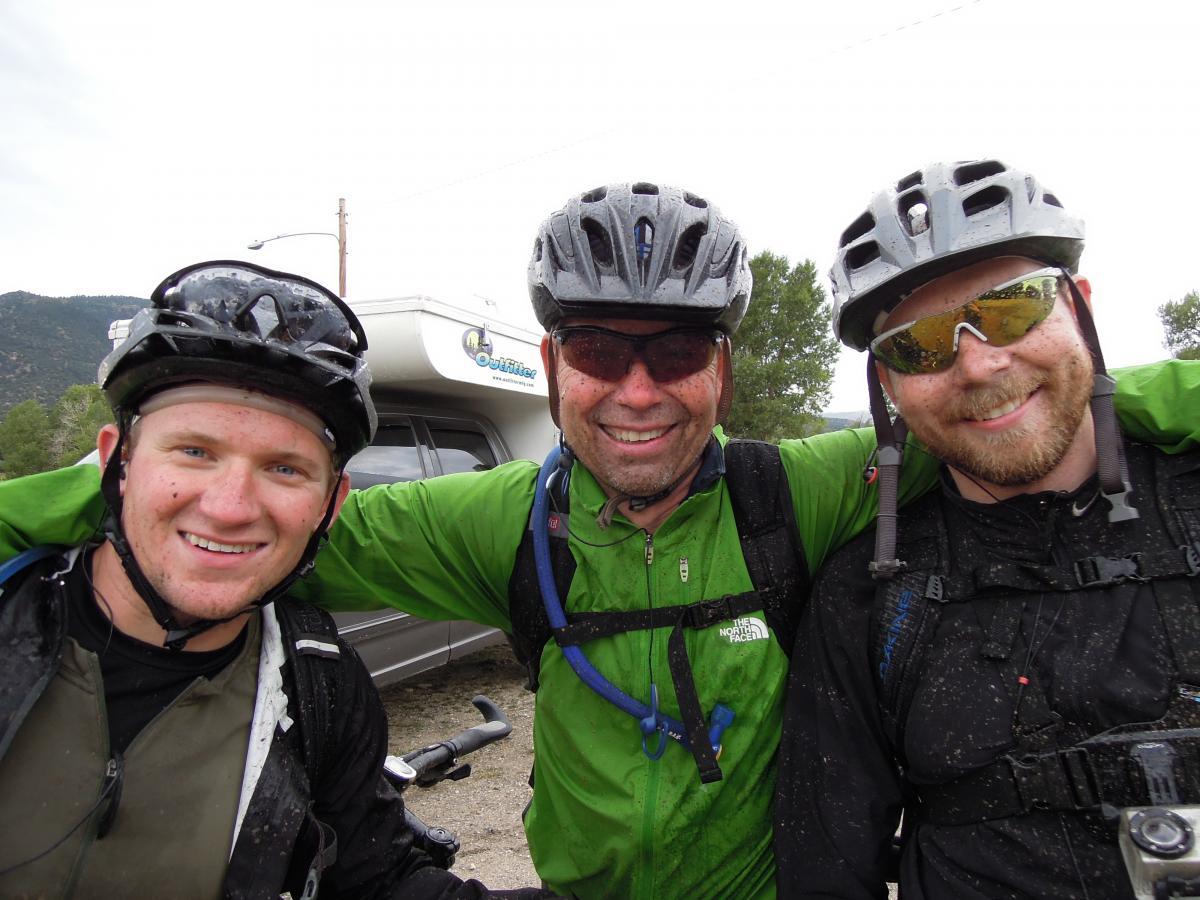 Three mountain bikers smiling and posing for a photo, standing close together with their arms around each other. They are wearing helmets and are slightly muddy from their ride. The background features a camper van and trees, with a cloudy sky above. Monarch Crest Trail mountain bike trail.
