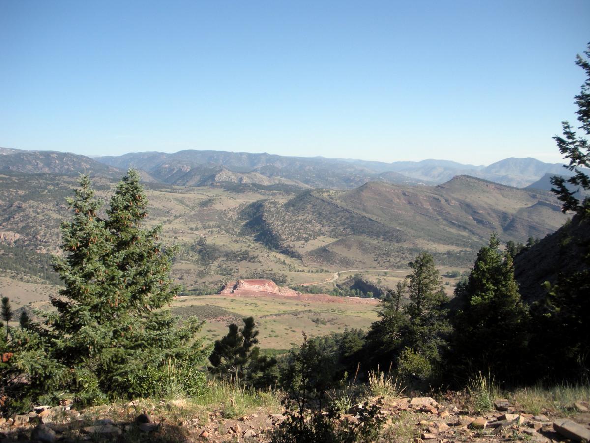 A scenic mountain landscape featuring rolling hills and valleys, with patches of greenery in the foreground. The sky is clear and blue, enhancing the natural beauty of the terrain. Heil Valley Ranch mountain bike trail.