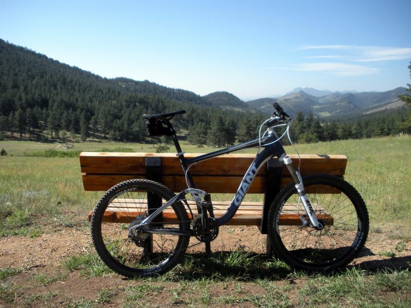 Giant Trance X3: A mountain bike leaning against a wooden bench, set against a scenic mountainous landscape with lush green hills and a clear blue sky.