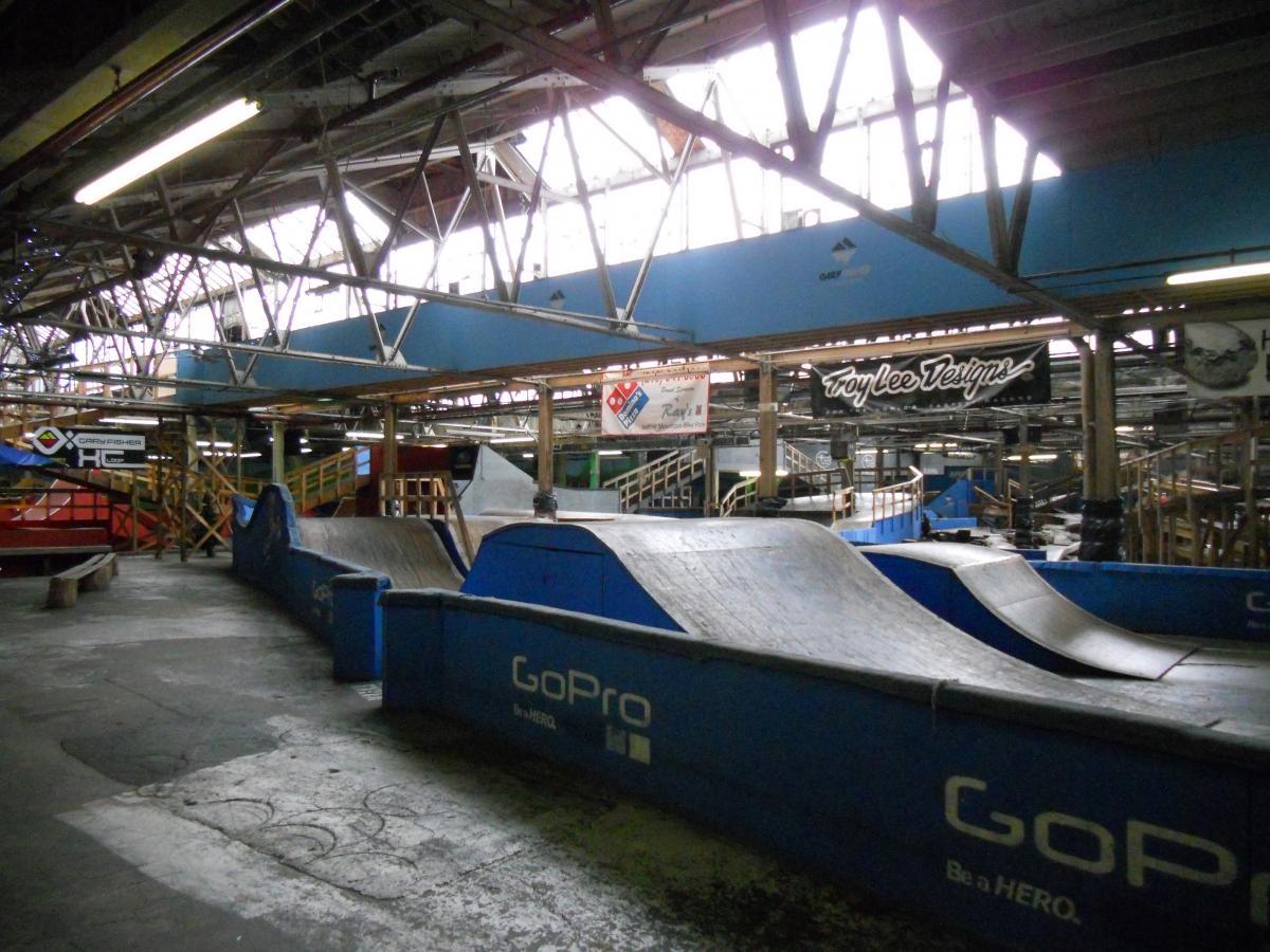 Indoor skatepark featuring ramps and a smooth concrete surface. The structure has high ceilings with visible metal beams and large windows, allowing natural light to illuminate the space. There are blue and red ramps, with branding for GoPro and Troy Lee Designs visible on the walls. The area appears empty, showcasing the design and layout of the park. Rays Indoor Mtb Park mountain bike trail.