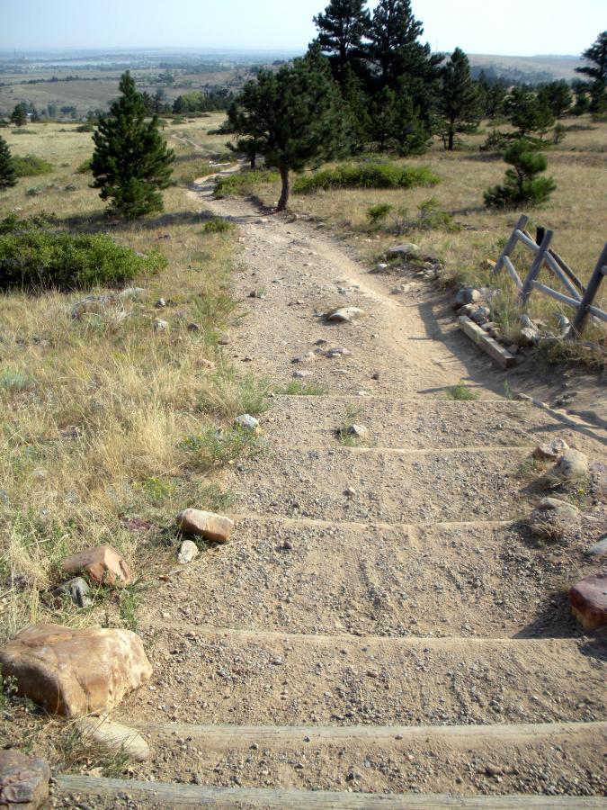 A dirt path descending through a grassy landscape, lined with rocks and surrounded by small trees, leading to a distant view of rolling hills and open sky. Greenbelt Plateau mountain bike trail.