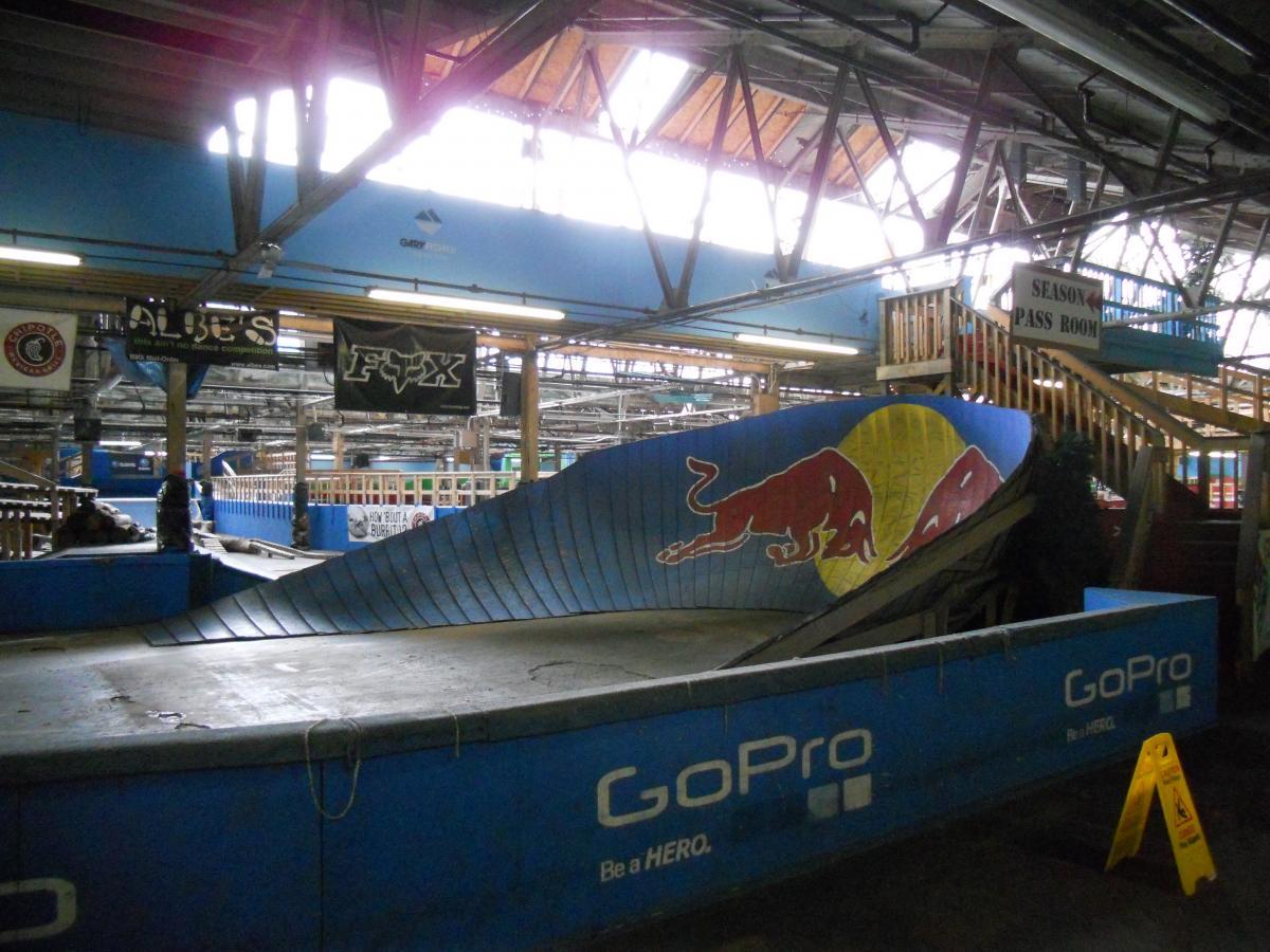 Indoor skate park featuring a curved ramp with a Red Bull logo, surrounded by banners and a seasonal pass room. The brightly lit space includes wooden beams and safety signs, with various skateboarding elements visible. Rays Indoor Mtb Park mountain bike trail.