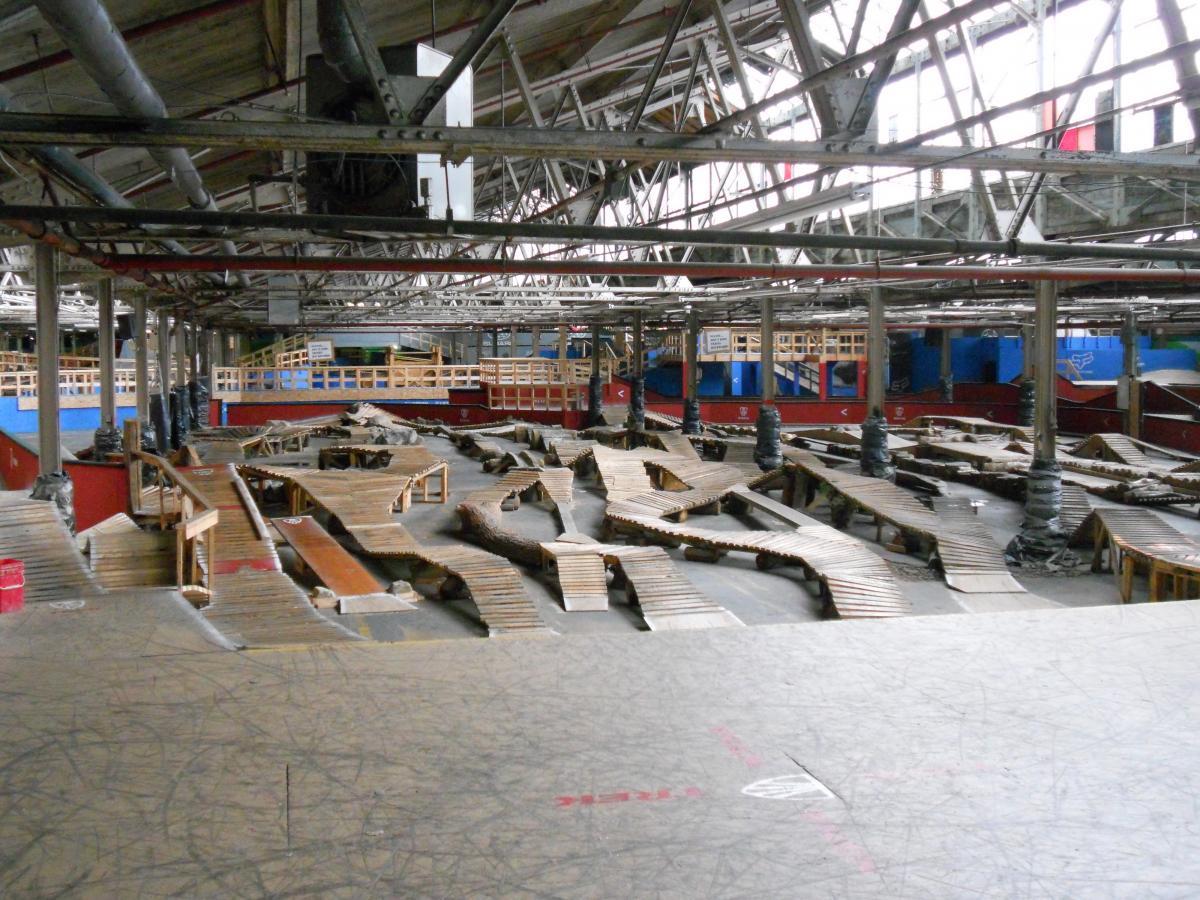 An expansive indoor skatepark featuring a complex layout of wooden ramps and trails, with a high ceiling supported by steel beams. The park includes elevated platforms, varying terrain designed for skateboarding or BMX biking, and colorful walls in the background. Rays Indoor Mtb Park mountain bike trail.