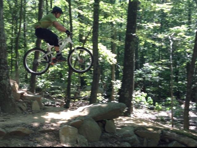 A mountain biker in a green shirt and helmet jumps off a rocky ledge in a forested area, surrounded by tall trees and greenery. The biker is airborne, showcasing a dynamic action shot. Vietnam Trails mountain bike trail.