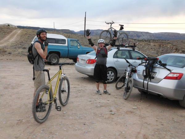 Two cyclists in a recreational area, one man smiling and waving excitedly while the other stands beside his yellow mountain bike, wearing a helmet. Several parked cars can be seen in the background, along with a scenic landscape of hills. Lunch Loops mountain bike trail.