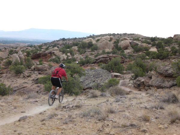 A person in a red shirt is riding a mountain bike on a dirt trail, navigating through a rugged landscape filled with rocks and sparse vegetation. Rolling hills and distant mountains are visible in the background under a cloudy sky. Lunch Loops mountain bike trail.