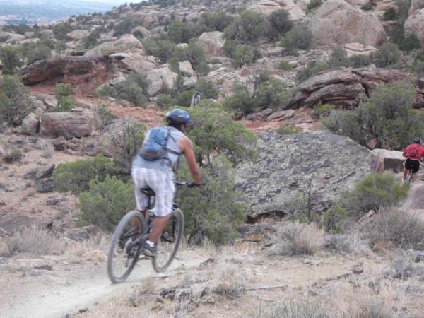 A mountain biker riding down a dirt trail in a hilly, rocky landscape, surrounded by shrubs and small trees. Another cyclist is visible in the background. The terrain appears rugged and natural, with large rocks and an expansive view. Lunch Loops mountain bike trail.