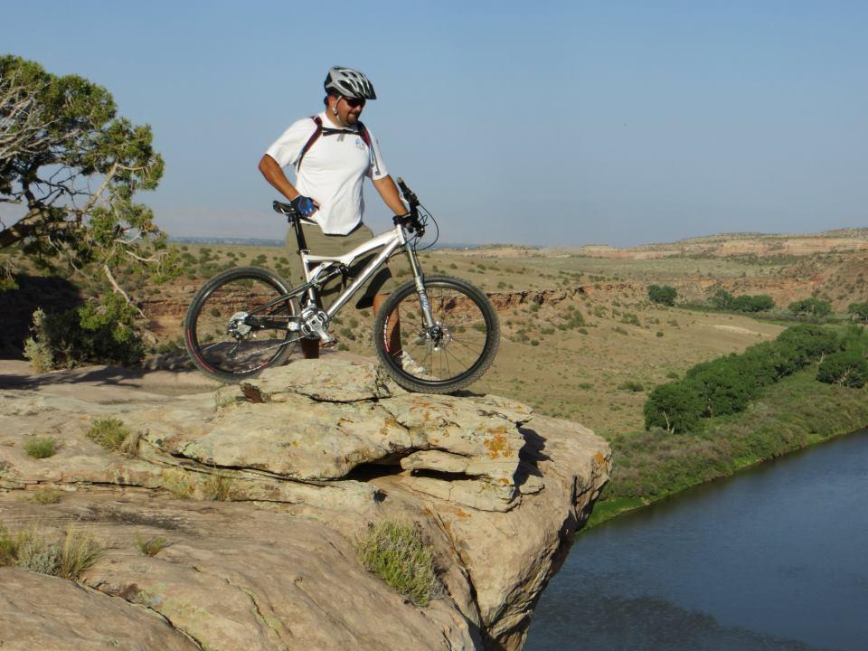 A mountain biker stands on the edge of a rocky outcrop, with a vast landscape of hills and a river visible below. The sky is clear and bright, suggesting a sunny day. The biker wears a helmet and sports attire, holding the bicycle while looking out over the scenic view. Kokopelli Area Trails mountain bike trail.