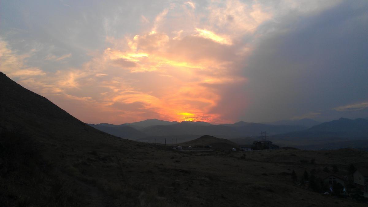 A serene sunset view over rolling hills and mountains, with vibrant hues of orange and pink in the sky, partially obscured by clouds. Silhouetted hills frame the scene, and a few structures can be seen in the foreground. North Table Mountain mountain bike trail.