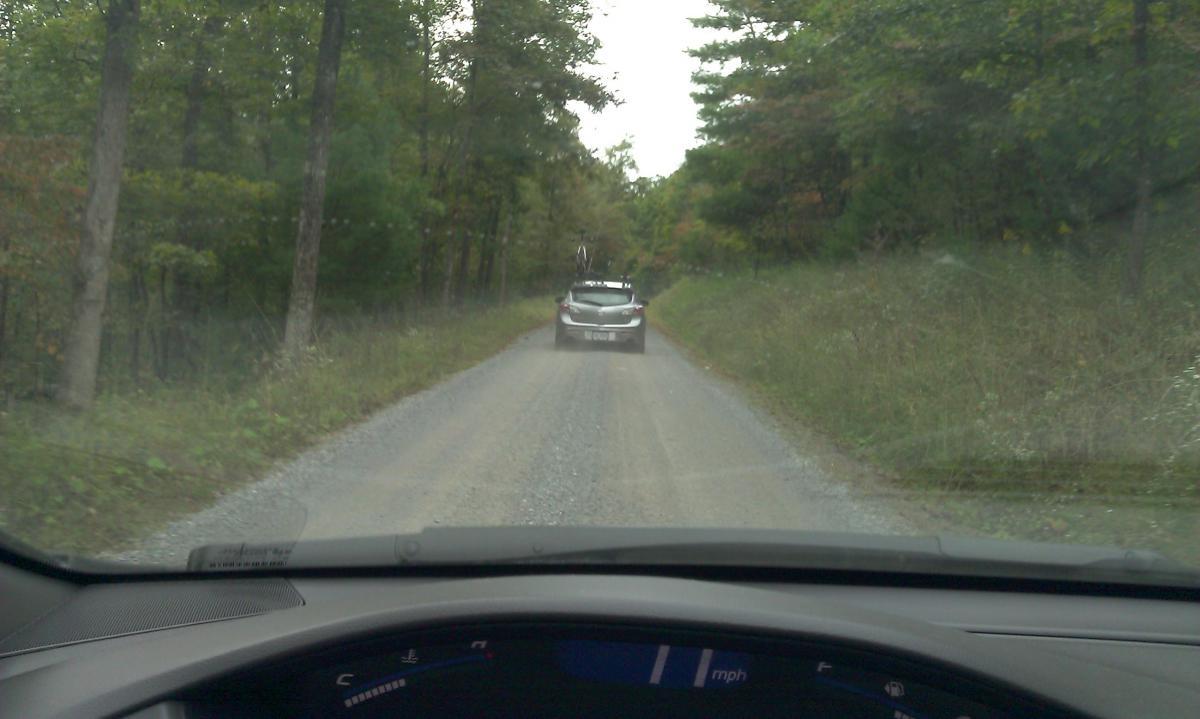 A view from inside a car looking down a gravel road, with a vehicle in front. The road is flanked by trees, showing signs of autumn foliage. The dashboard displays a speedometer with a reading of mph. Reddish Knob mountain bike trail.