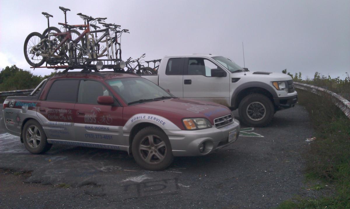 Two vehicles parked at a scenic overlook. A red and silver car with a bike rack on top holds several bicycles, while a white truck is parked beside it. The background features greenery and a cloudy sky. The ground is marked with chalk graffiti. Reddish Knob mountain bike trail.