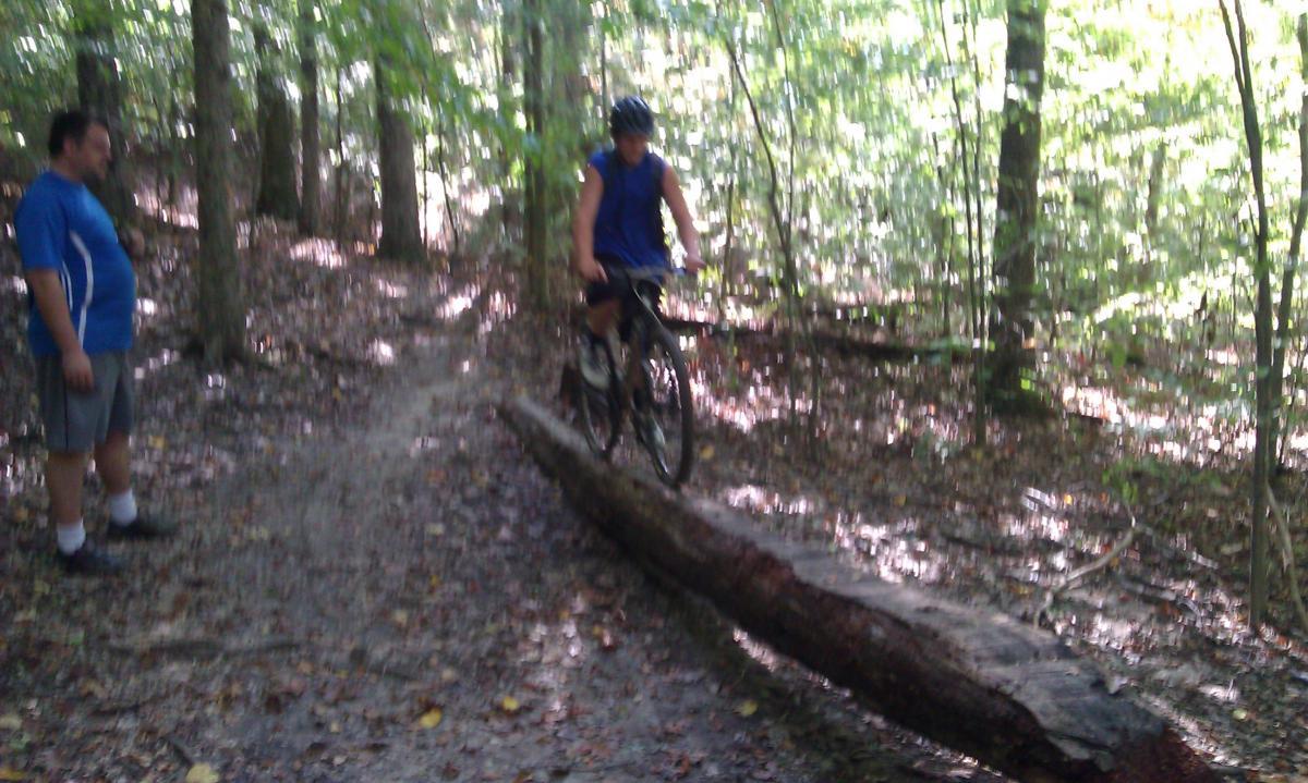 A mountain biker in a blue tank top rides over a fallen log on a forest trail while another person watches nearby. The scene is surrounded by trees with dappled sunlight filtering through the leaves. Schaeffer Farms mountain bike trail.