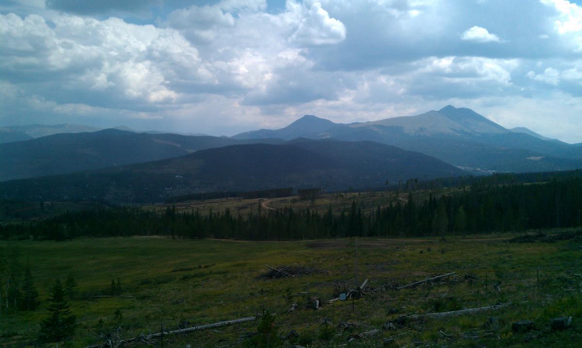 A panoramic view of a mountain range under a partly cloudy sky, featuring rolling hills and a grassy foreground with scattered trees. A winding dirt road can be seen leading through the landscape, creating a sense of depth and exploration in this serene natural setting. Peaks Trail mountain bike trail.