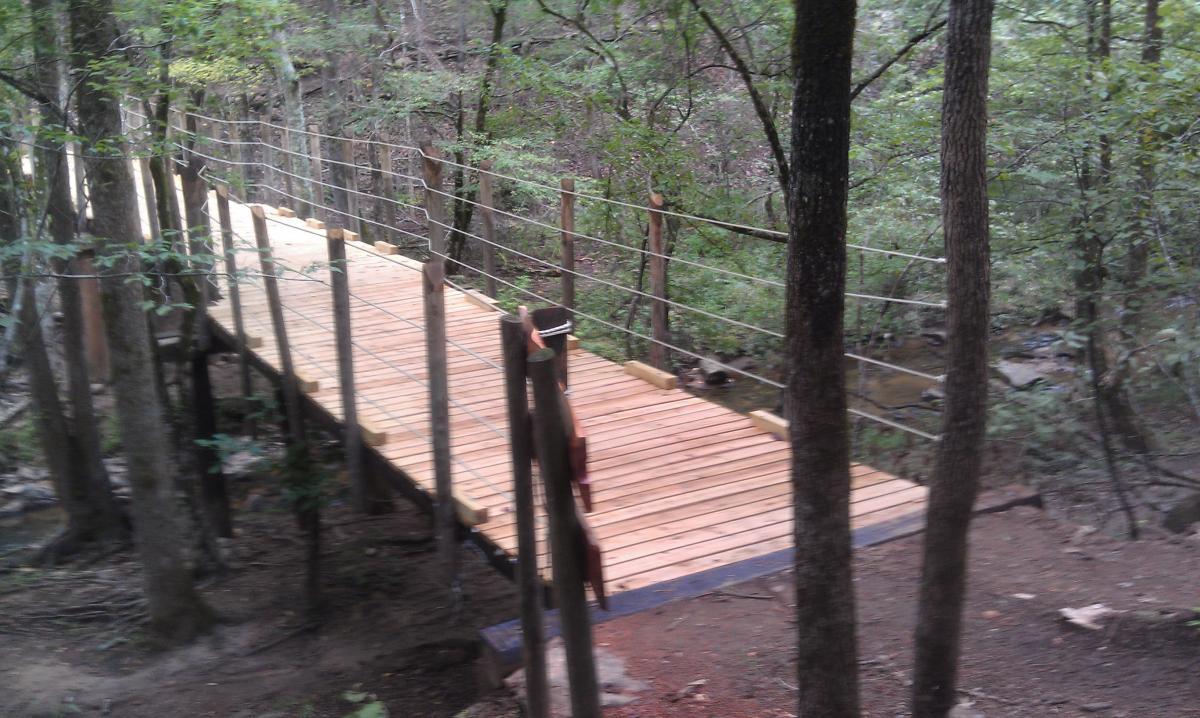 A wooden footbridge with rope railings spans a small creek, surrounded by dense trees and greenery in a forested area. Chewacla State Park mountain bike trail.