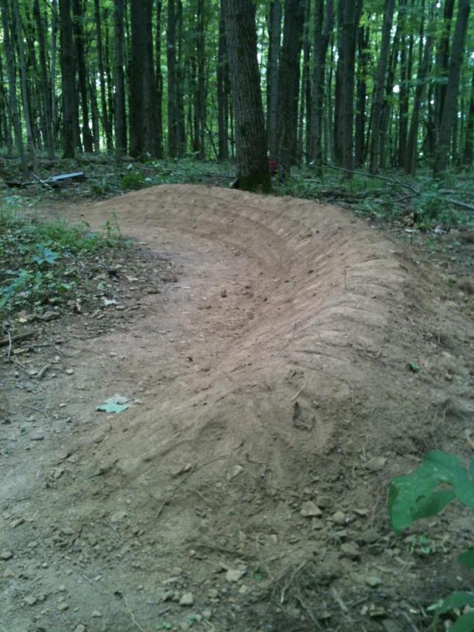 A curved dirt bike trail located in a wooded area, featuring a smooth, packed surface and surrounded by trees and greenery. The trail is gently sloping and looks designed for biking or recreational use. Nockamixon State Park mountain bike trail.