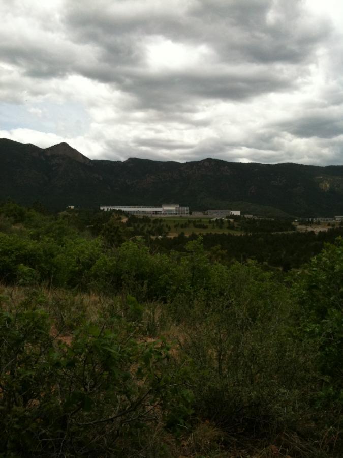 A view of a mountainous landscape under a cloudy sky, featuring a building nestled among greenery and trees in the foreground. The mountains rise in the background, creating a serene and natural setting. Falcon Trail mountain bike trail.