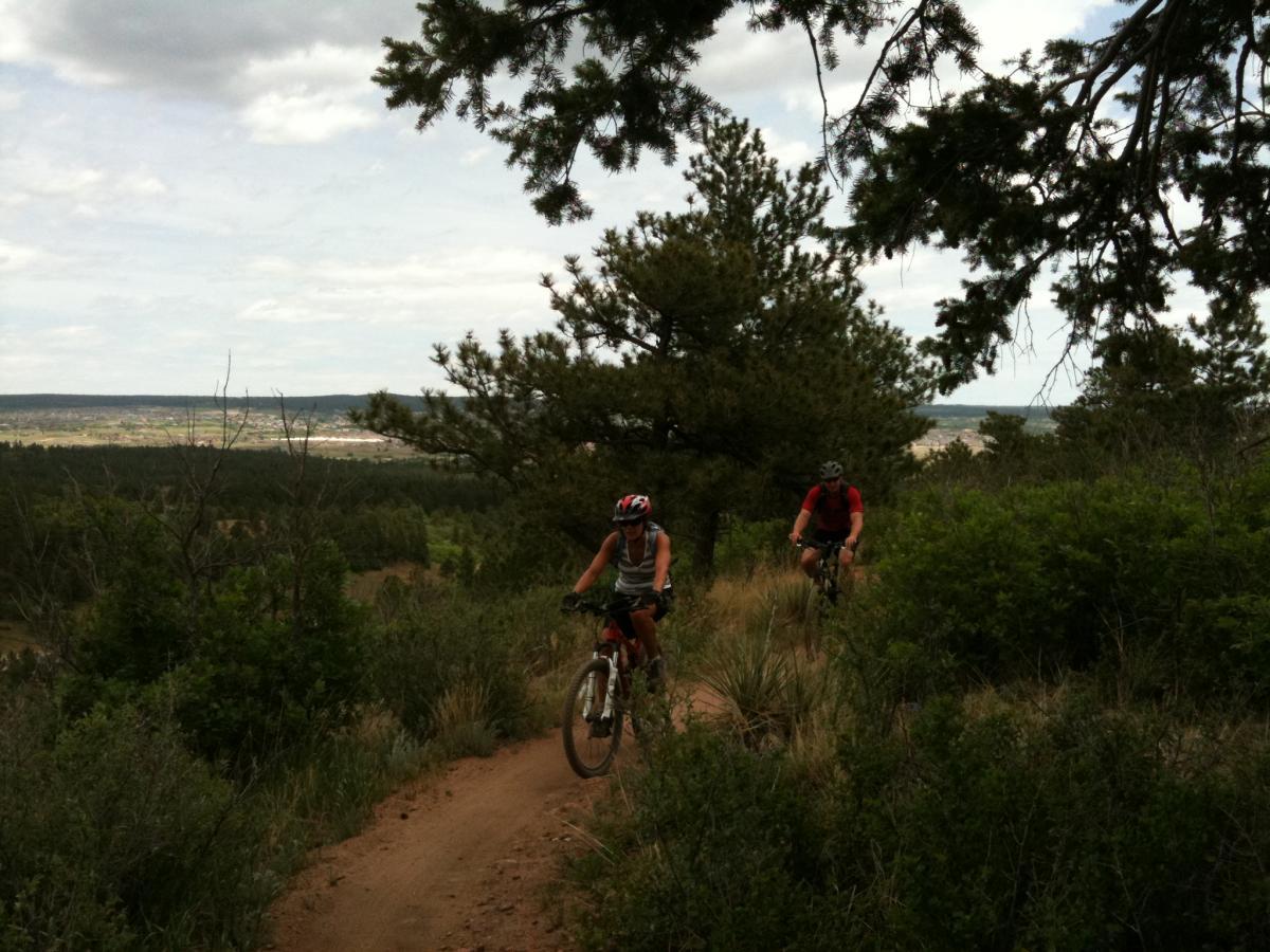Two cyclists navigating a dirt trail in a mountainous area, surrounded by trees and greenery. The landscape features rolling hills and a distant view of a valley under a cloudy sky. Falcon Trail mountain bike trail.