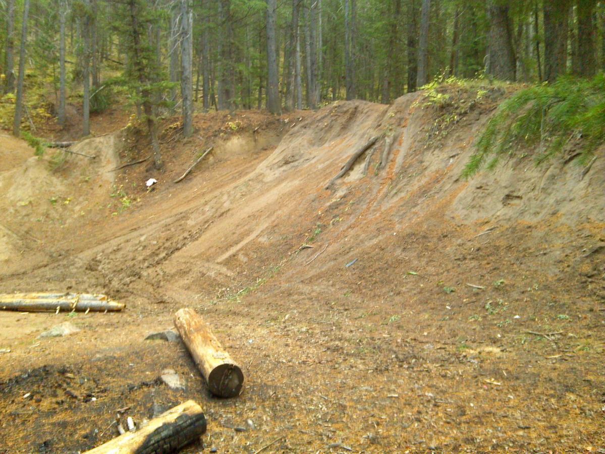 A sandy clearing in a forest, featuring a steep, sloped embankment. The area is surrounded by tall trees with sparse foliage, and some fallen logs are scattered on the ground. The soil appears disturbed, indicating possible erosion or previous activity in the area. Waterfall Loop mountain bike trail.