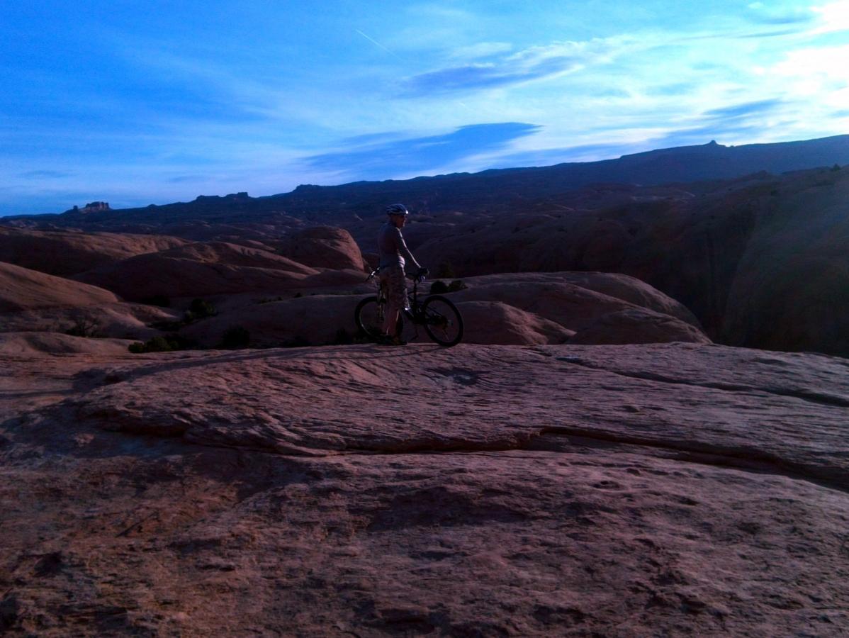 A person standing next to a mountain bike on a rocky terrain, with a scenic landscape of hills and blue sky in the background during sunset. Slickrock mountain bike trail.