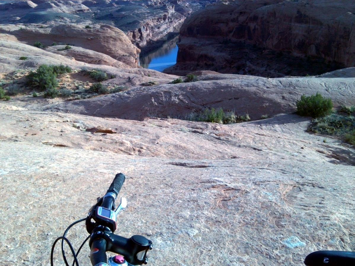A mountain bike closely positioned in the foreground, with a rocky landscape and a river visible in the background. The scene captures a natural canyon with steep rock formations and sparse vegetation under a clear sky. Slickrock mountain bike trail.