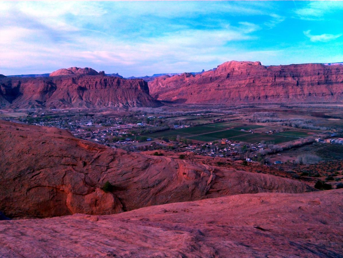 A panoramic view of a canyon landscape at dusk, showcasing red rock formations and rolling hills. In the foreground, rugged terrain leads to a valley below, where a small town with scattered houses and agricultural fields is visible. The sky above is partly cloudy, transitioning to soft hues of blue and pink as the sun sets. Slickrock mountain bike trail.