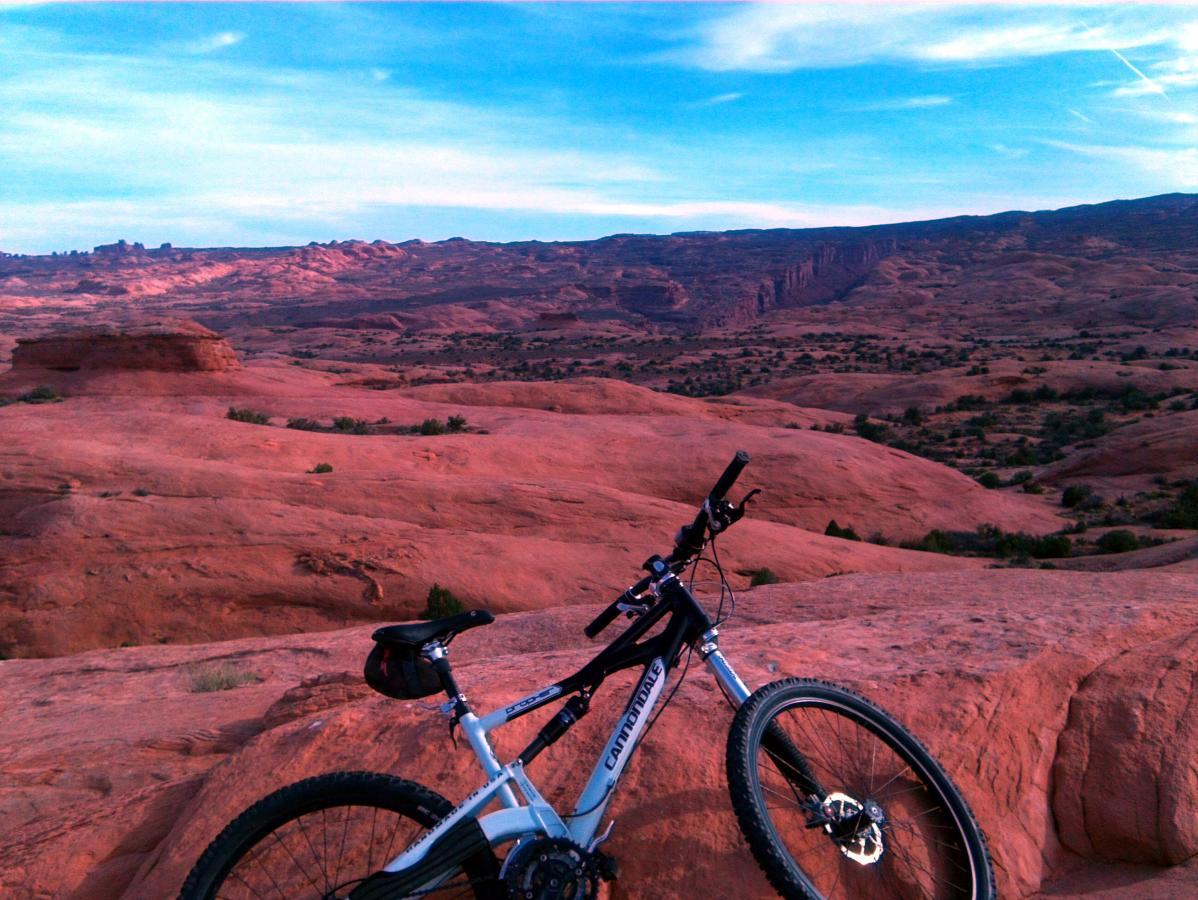 A mountain bike resting on red rock formations, with a vast desert landscape and distant mountains under a blue sky. Slickrock mountain bike trail.