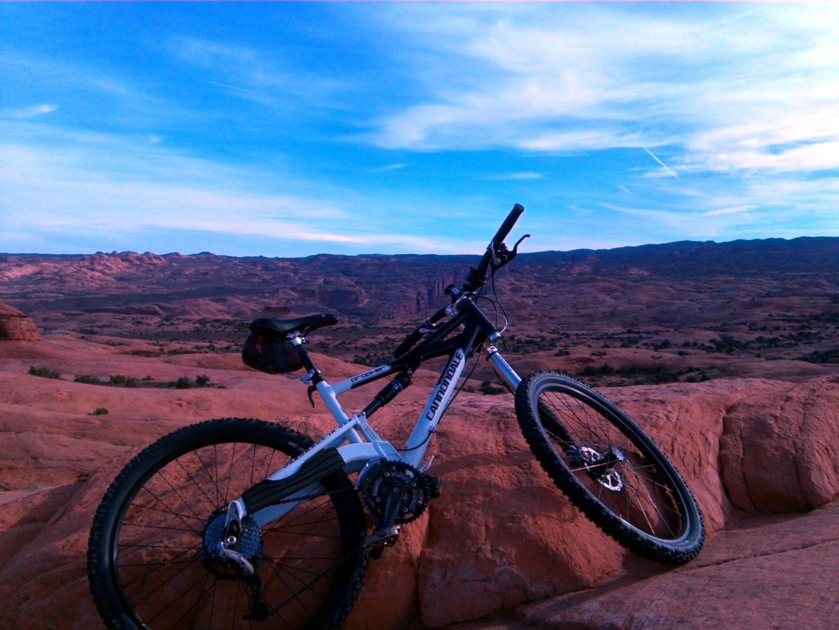 A mountain bike resting on a rocky surface with a vast desert landscape in the background under a blue sky. The scene captures the natural beauty and rugged terrain, ideal for outdoor biking adventures. Slickrock mountain bike trail.