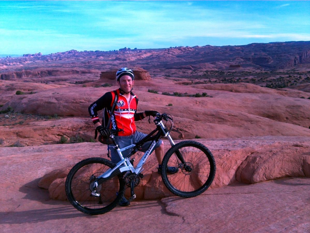 A mountain biker stands proudly next to his bike on a rocky landscape, with a vast desert background under a blue sky. He is dressed in riding gear, featuring a red and black jersey. The scene showcases rugged terrain and distant mountains, highlighting the adventurous spirit of outdoor cycling. Slickrock mountain bike trail.
