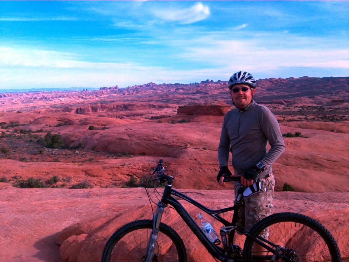A person wearing a helmet and sunglasses stands next to a mountain bike on a rocky, red landscape. The background features rolling hills and a clear blue sky, highlighting a scenic outdoor adventure setting. Slickrock mountain bike trail.