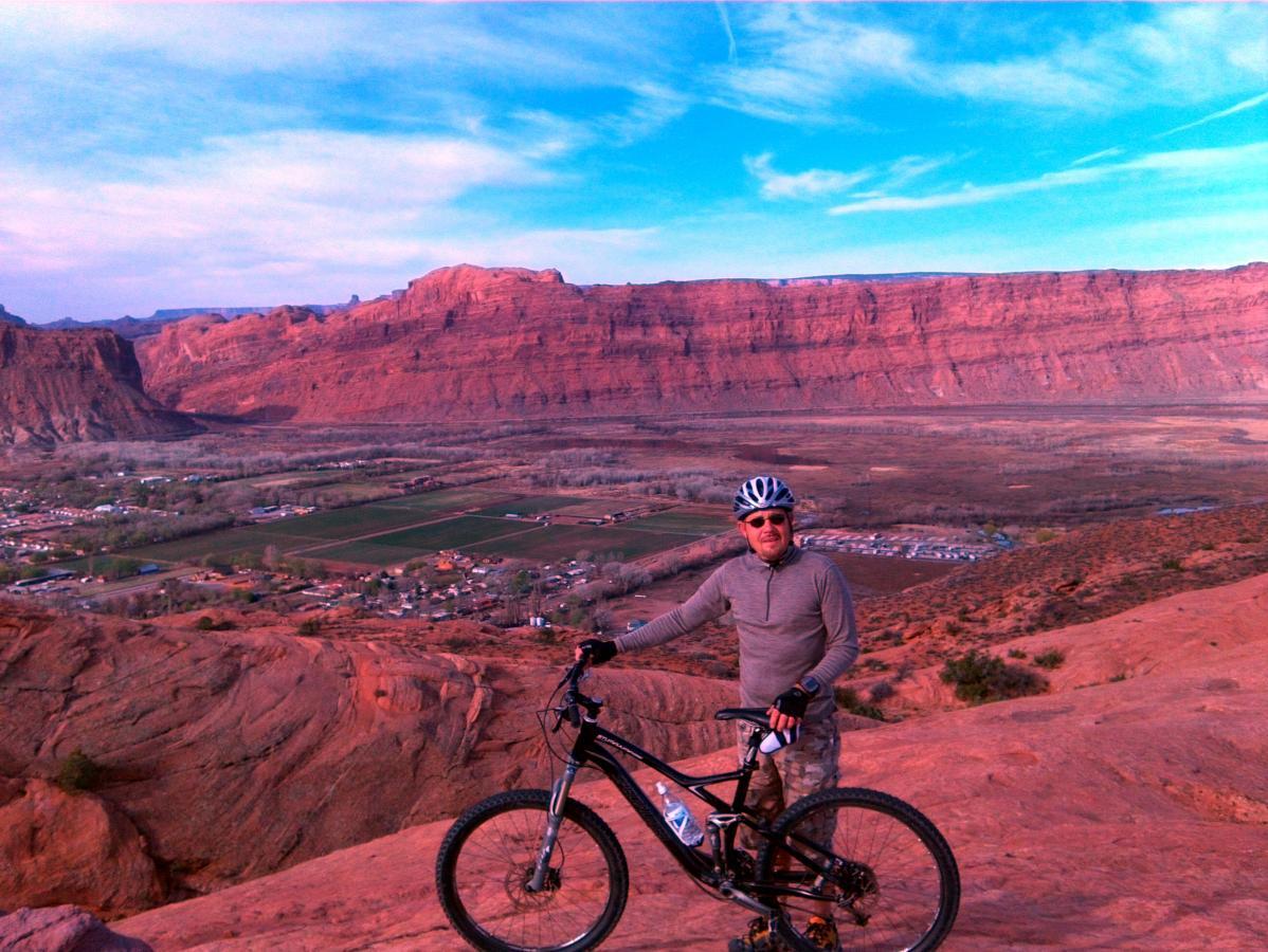 A mountain biker stands on a rocky ledge overlooking a scenic valley, surrounded by red rock formations under a blue sky. The bike rests beside him, and the landscape features a mix of greenery and residential areas in the distance. Slickrock mountain bike trail.