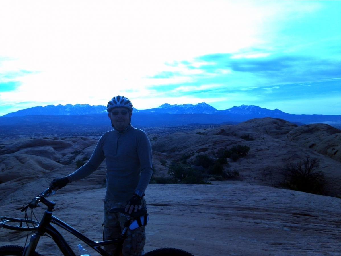 A mountain biker wearing a helmet and sunglasses stands beside his bike, overlooking a dramatic landscape of rolling hills and distant snow-capped mountains under a blue sky. The scene captures the spirit of adventure and the beauty of nature. Slickrock mountain bike trail.