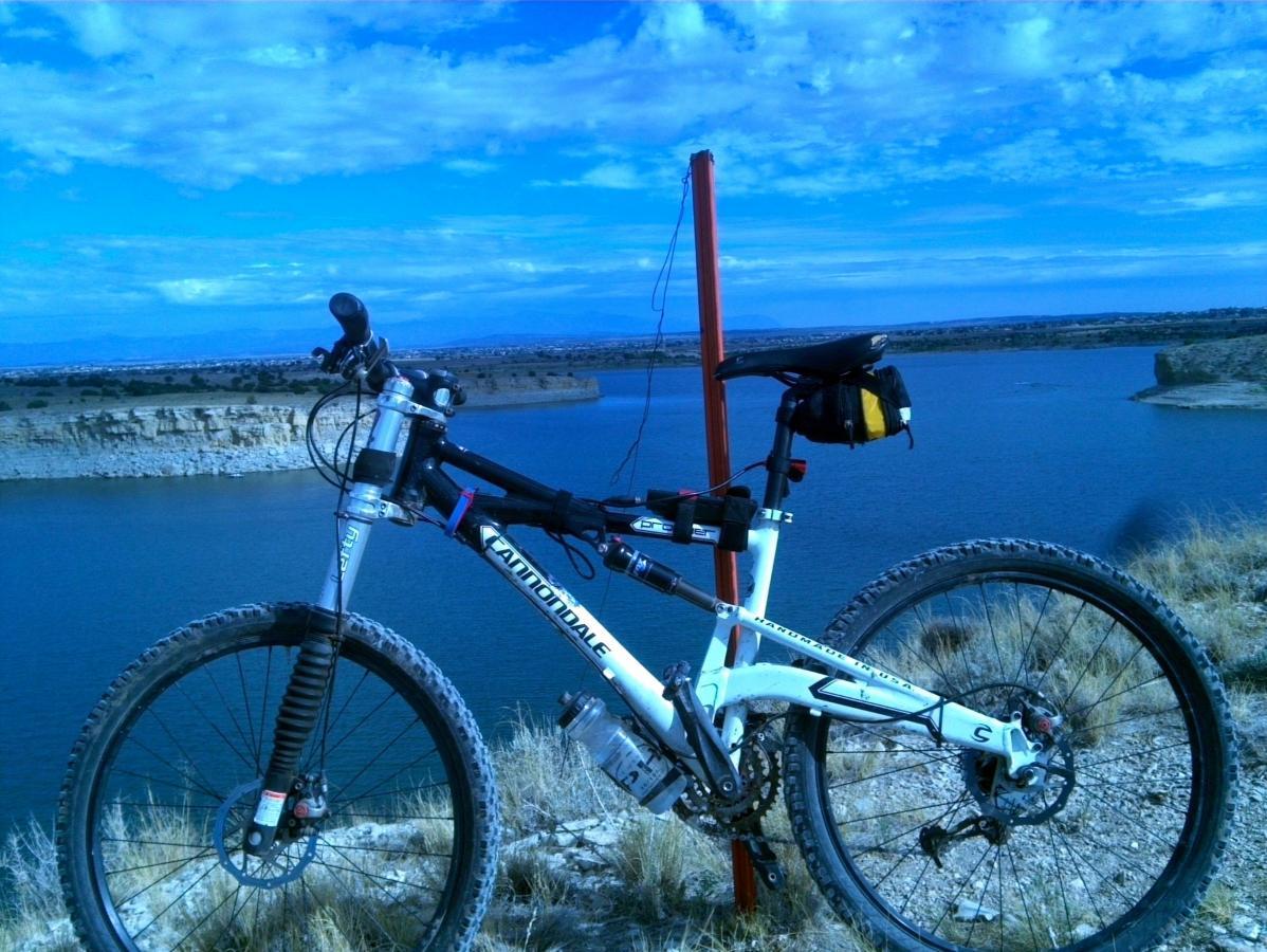 A mountain bike parked on a rocky ledge overlooking a body of water, with a scenic view of the sky and hills in the background. The bike features a white frame and is equipped with a small black bag attached to the seat. South Shore Lake Pueblo mountain bike trail.