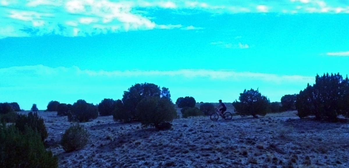 A silhouette of a person riding a bicycle across a serene, open landscape with low shrubs under a bright blue sky filled with wispy clouds. South Shore Lake Pueblo mountain bike trail.