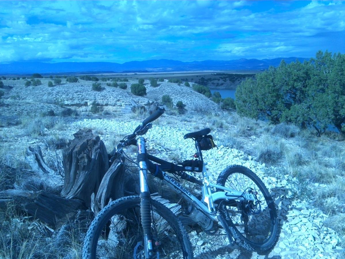 A mountain bike resting on a rocky path overlooking a scenic landscape with rolling hills, sparse vegetation, and a river in the distance under a blue sky filled with clouds. South Shore Lake Pueblo mountain bike trail.