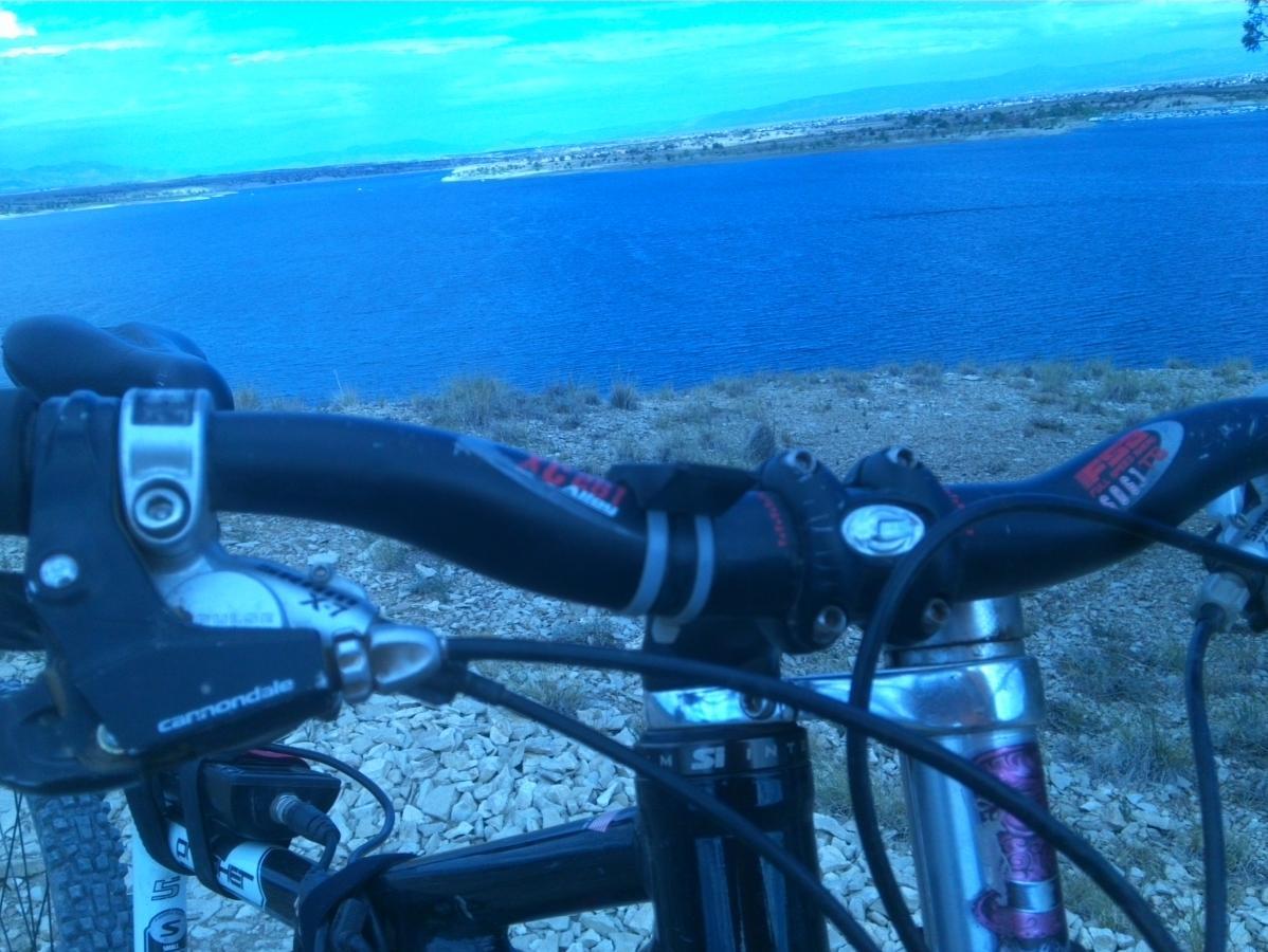 A close-up view of a mountain bike's handlebars, with a lake and a clear blue sky in the background. The image captures the bike's grips and gear shifters against the serene water, suggesting an outdoor cycling adventure. South Shore Lake Pueblo mountain bike trail.