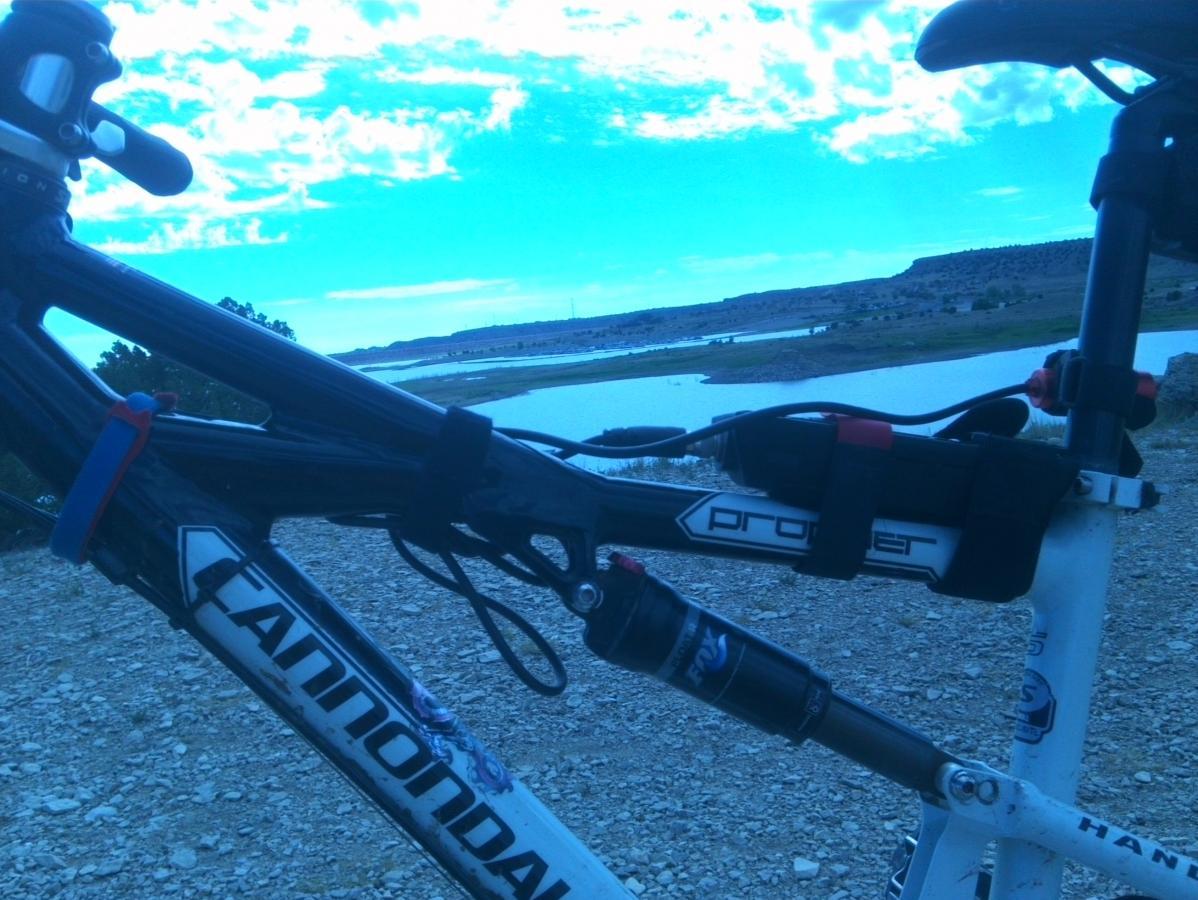 A close-up view of a black Cannondale mountain bike frame with a water bottle holder and suspension system, set against a backdrop of a serene lake and blue sky with scattered clouds. The ground is rocky, indicating an outdoor setting, possibly on a trail or near a biking path. South Shore Lake Pueblo mountain bike trail.