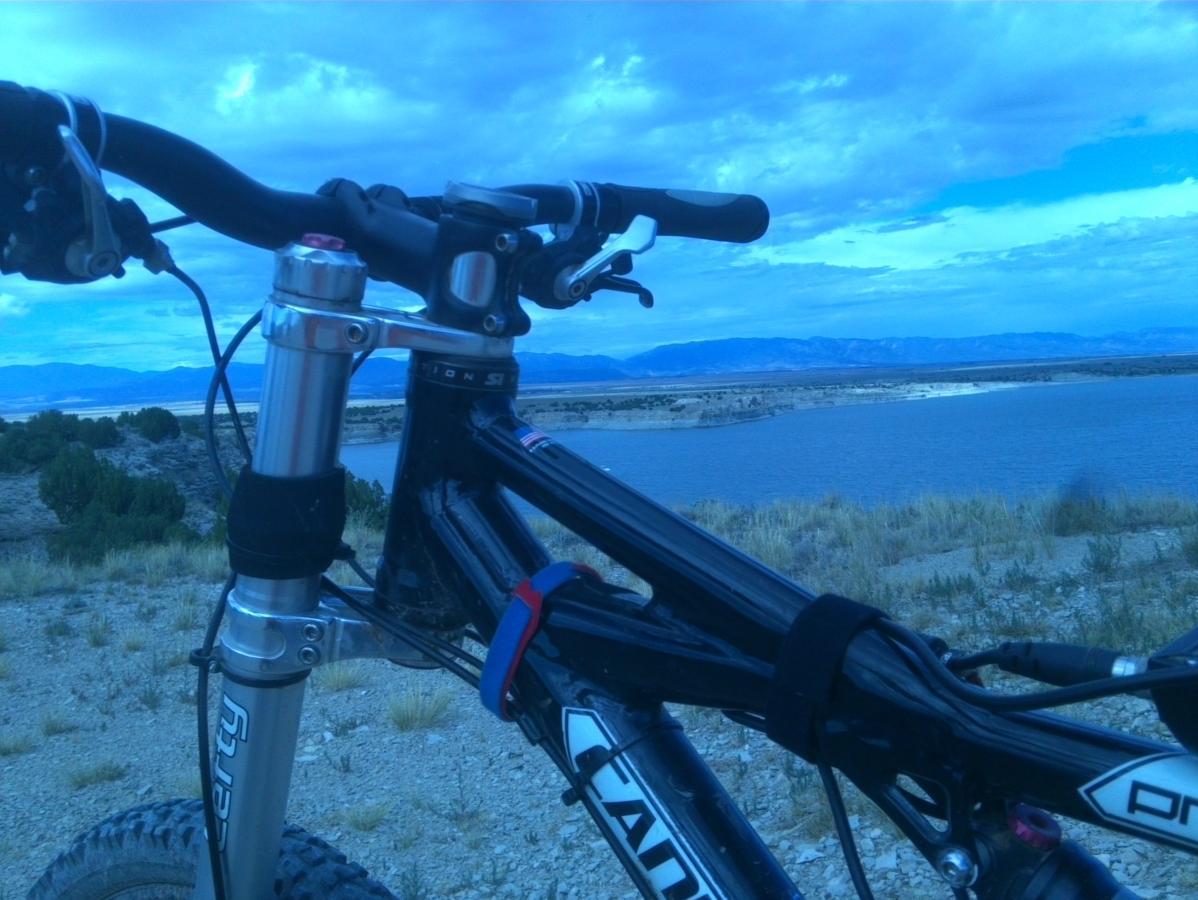 Close-up view of a mountain bike's handlebars and frame, with a scenic landscape in the background featuring a body of water and distant mountains under a cloudy blue sky. The bike is positioned on rocky terrain with sparse vegetation. South Shore Lake Pueblo mountain bike trail.