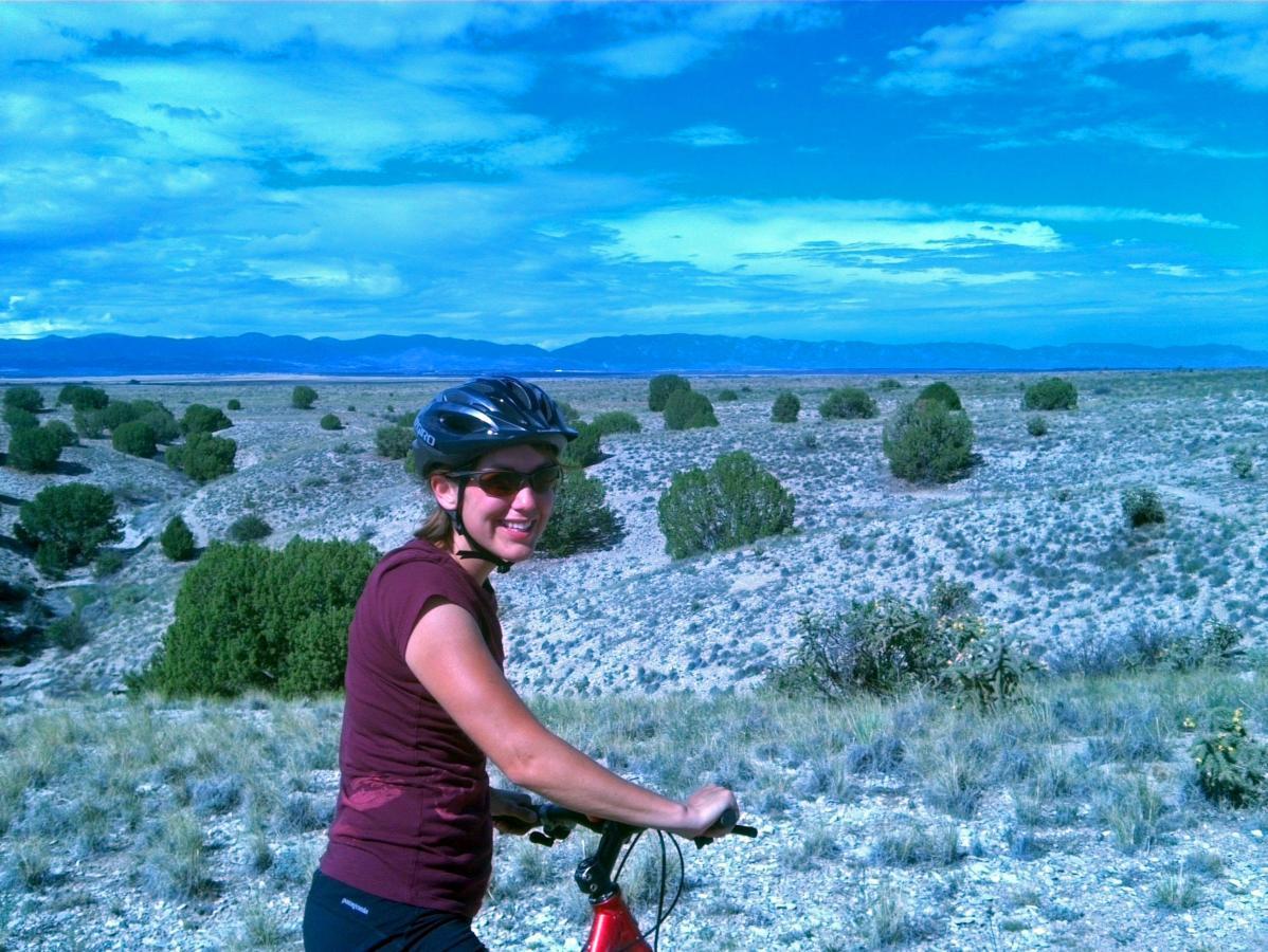 A woman in a cycling helmet and sunglasses smiles while riding her bike on a gravel trail, with a vast landscape of shrubs and rolling hills in the background under a blue sky. South Shore Lake Pueblo mountain bike trail.