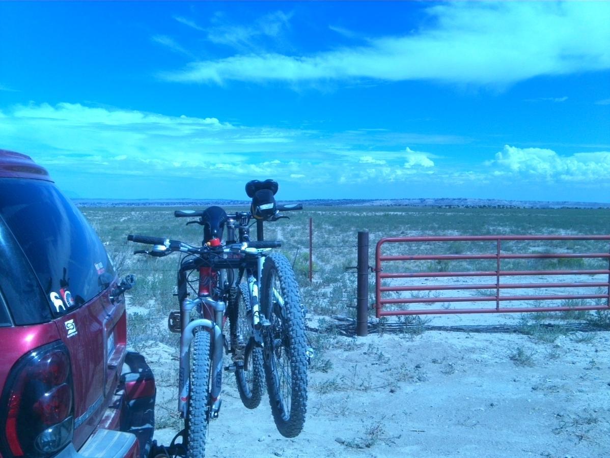 A red vehicle with bicycles mounted on a rack is parked near a wide-open landscape under a bright blue sky with scattered clouds. In the background, a red gate marks the entrance to a grassy area. South Shore Lake Pueblo mountain bike trail.