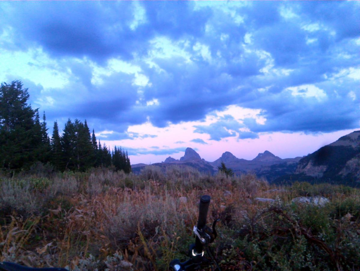 A panoramic view of a mountainous landscape during twilight, featuring rolling hills and peaks under a cloudy sky. In the foreground, there are patches of grass and shrubs, along with the handlebar of a bicycle partially visible on the right. The colors of the sky transition from blue to soft pink as the sun sets in the background. Pine trees line the left side of the image. Mill Creek mountain bike trail.