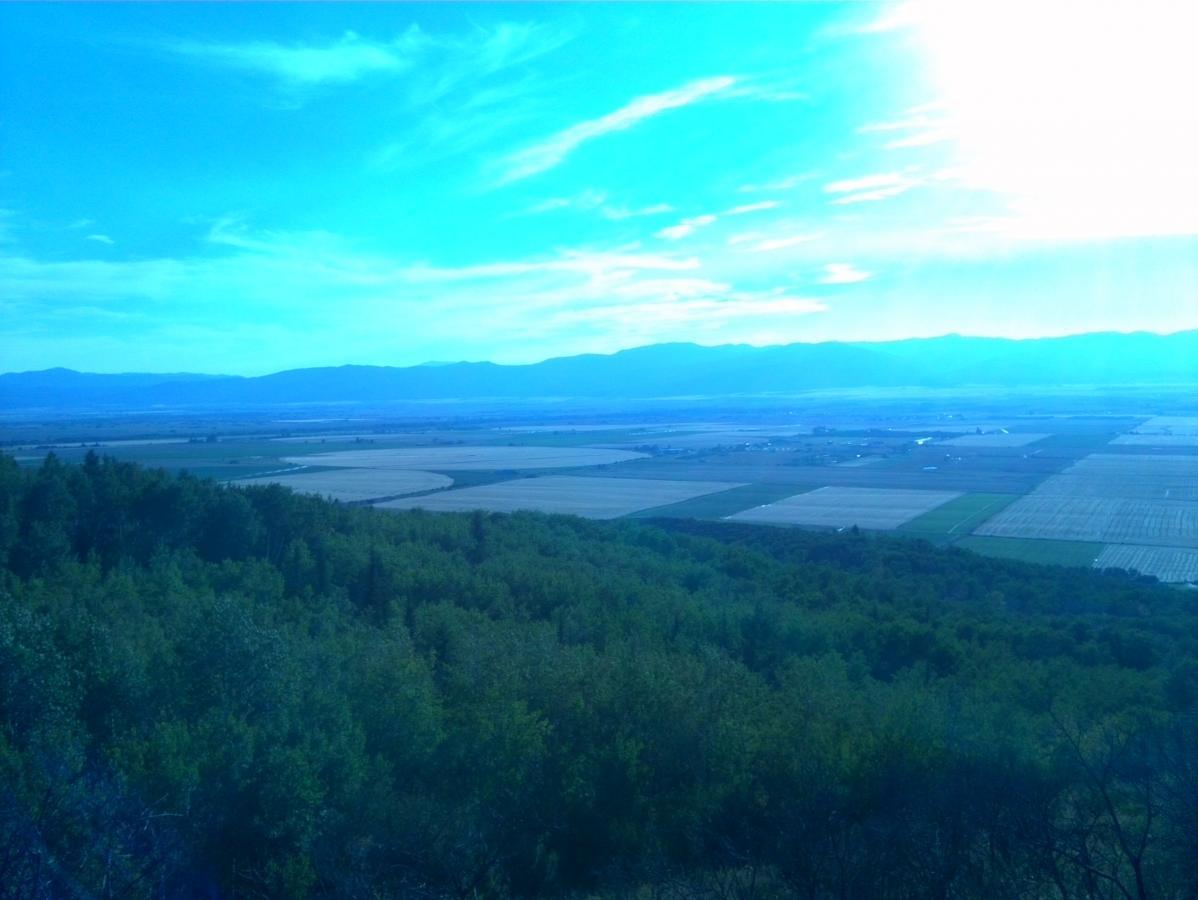 A panoramic view of a lush green landscape with fields and patches of cultivated land extending into the distance. The scene is set against a backdrop of mountains under a bright blue sky with scattered clouds, creating a tranquil and picturesque rural environment. Aspen Trail mountain bike trail.