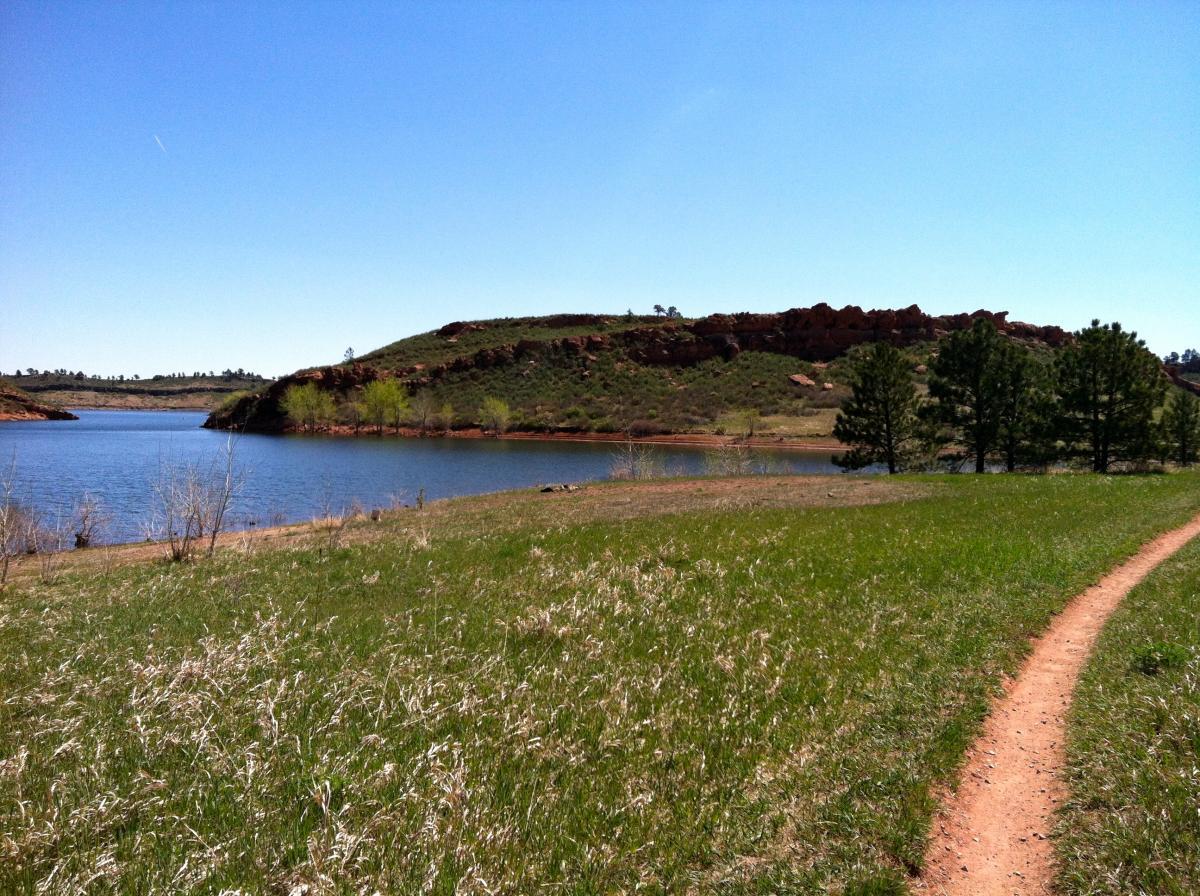 A serene landscape featuring a calm lake bordered by green grass and scattered shrubs. In the background, rolling hills are visible under a clear blue sky. A dirt path meanders along the water's edge, inviting exploration. Lory State Park mountain bike trail.