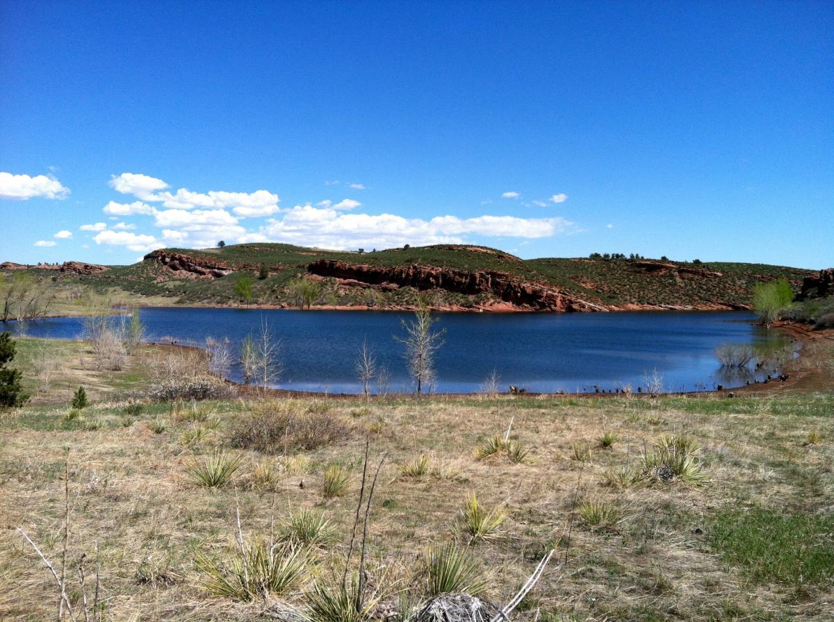 A scenic landscape featuring a calm lake surrounded by grassy fields and hills. The clear blue sky is dotted with a few white clouds, and the shoreline is lined with sparse vegetation, including small trees and shrubs. The distant hills display a mixture of greenery and reddish rock formations, creating a tranquil natural setting. Lory State Park mountain bike trail.
