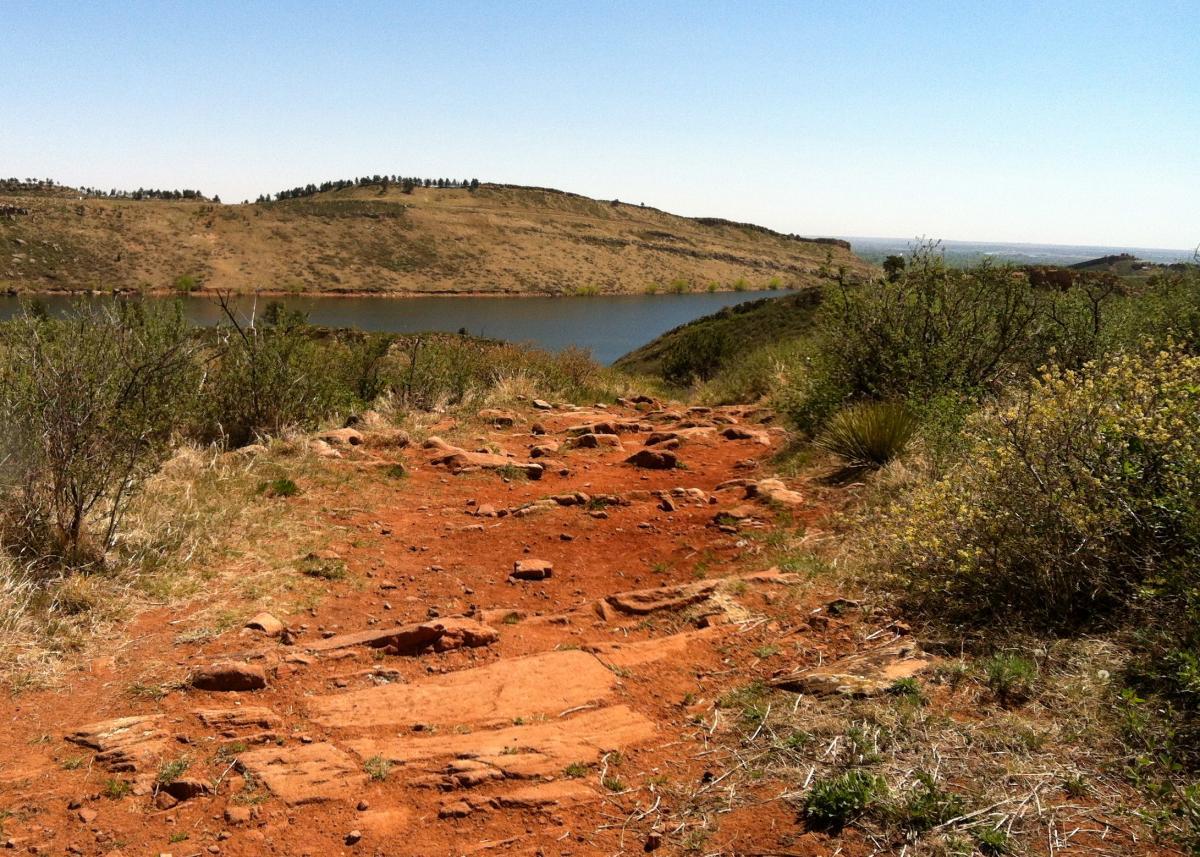 A dirt path with reddish soil leads towards a calm lake surrounded by hills. Lush green foliage and small bushes flank the sides of the trail, while a clear blue sky stretches above. In the background, rolling hills are visible, creating a serene natural landscape. Lory State Park mountain bike trail.