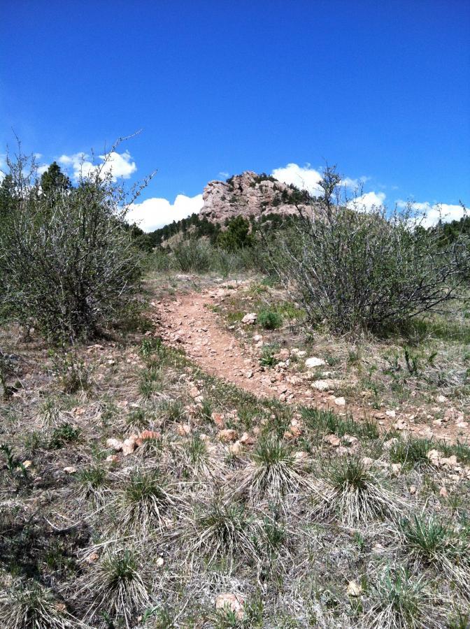 A dirt trail winding through a rugged landscape, surrounded by sparse bushes and grass, leading towards a rocky formation under a clear blue sky with a few clouds. Lory State Park mountain bike trail.