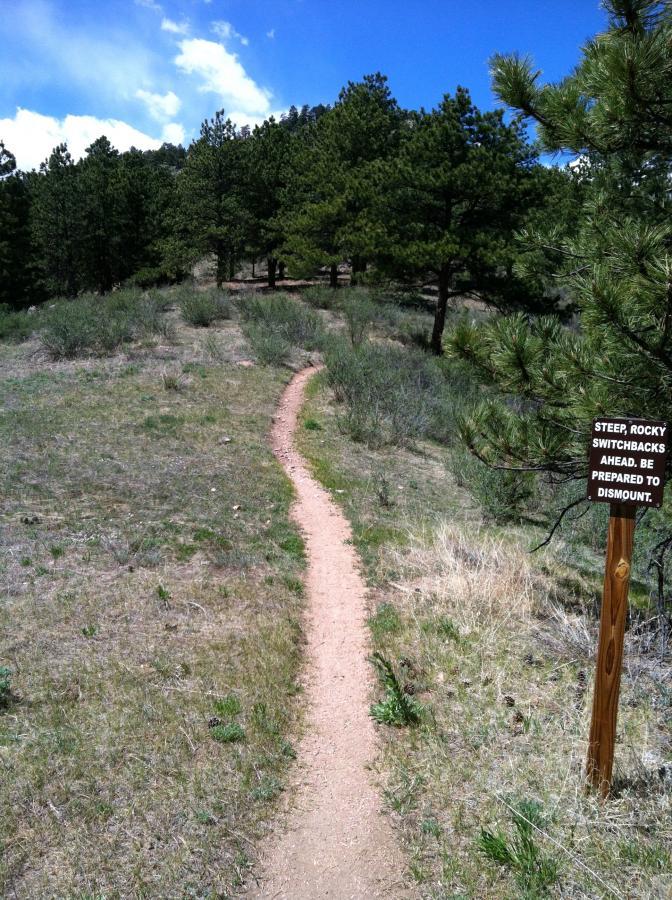 A dirt hiking path winding through a grassy area lined with trees, with a warning sign on the right that reads, "Steep, Rocky Switchbacks Ahead. Be Prepared to Dismount." The scene is set under a blue sky with scattered clouds. Lory State Park mountain bike trail.